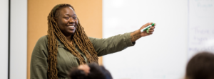 A teacher stands at the front of a classroom with a marker in hand, smiling.