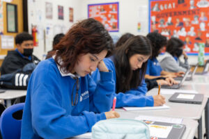 Students sit at desks, concentrating on their work with pencils in hand and laptops nearby.