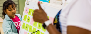 A student points at letters on a board while their teacher gives them a thumbs-up.