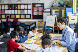 A teacher sits a table surrounded by four students, all looking at the same book.