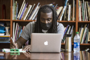 Teacher works at his laptop with a bookshelf behind him.