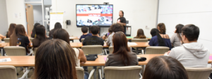 Shown from behind, a classroom full of students listen in a presenter at the front of the room.