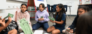 Students seated on a classroom floor hold green play money, with worksheets spread out in front of them.