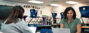 Two women sit across the table from each other working on laptops.