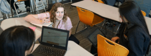 An adult with an open notebook crouches next to two students working on laptops.