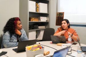 Two women seated at a table with open laptops engaged in discussion.