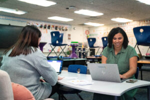 Two women sit across the table from each other working on laptops.