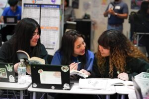 Three students sit side-by-side, reading open books and engaging in discussion.